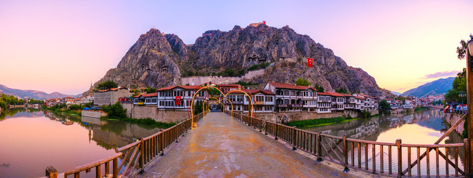 Twilight view of Amasya, Turkey. Traditional Ottoman houses along Yesilirmak River below ancient Pontic King Rock Tombs. A glowing bridge spans the river under a colorful sky. Historic heritage site.