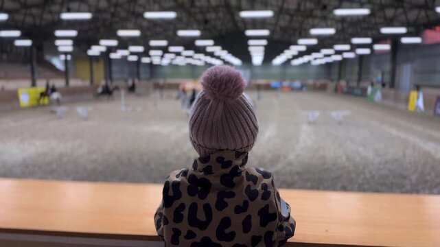 Young spectator sitting on a spectator stand, intently watching a horse riding lesson in a large indoor arena. Little girl with a pom pom hat enjoying the equestrian sport competition