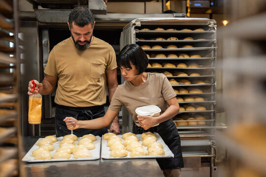Bakers glazing fresh buns on trays in artisan bakery kitchen
