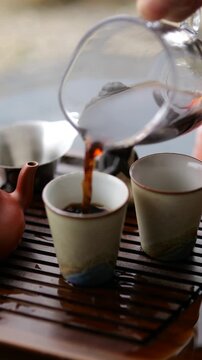 Close up of a tea master pouring hot, dark puerh tea from a glass pitcher into ceramic cups during a traditional gongfu cha ceremony, showcasing the ancient art of tea drinking