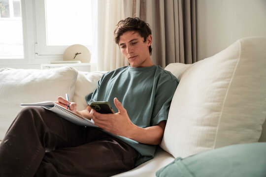 Young man taking notes while using smartphone on sofa
