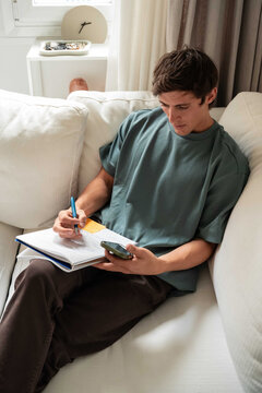 Young man taking notes on sofa with phone at home