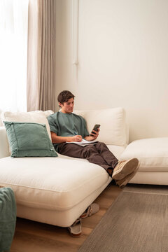 Young man using smartphone and notebook on sofa at home