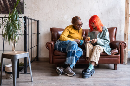 Women friends on sofa sharing smartphone and laughing at home