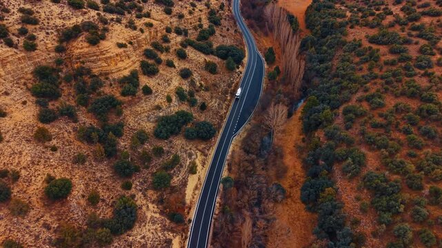 Drone view of winding rural road in Spain. Top-down drone view of black asphalt road through dry ocher terrain with shrubs and one vehicle.