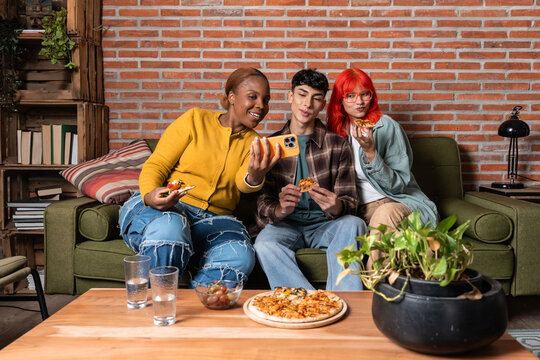 Friends taking selfie while eating pizza on cozy sofa