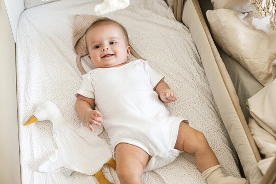 Cute little baby lying in crib with sweet toy and boho mobile. Bedtime. Adorable infant portrait in bassinet and smiling, space for text. Childhood and childcare.