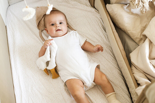 Cute little baby lying in crib with sweet toy and boho mobile. Bedtime. Adorable infant portrait in bassinet, space for text. Childhood and childcare.