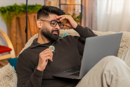 Indian man at home reacts to Bitcoin BTC drop, closing laptop and holding coin with unhappy look. Arabian guy on sofa expresses disappointment and stress, emotionally affected by falling crypto value