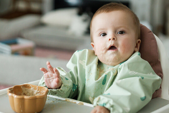 Cute little baby try to eat solid food in child chair. Complementary food. Adorable happy messy and funny infant portrait in porridge. Feeding after baby 6 months