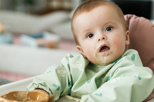 Cute little baby try to eat solid food in child chair. Complementary food. Adorable messy and funny infant portrait in porridge. Feeding after baby 6 months