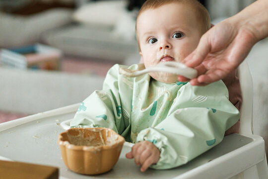 Cute little baby try to eat solid food in child chair. Complementary food. Adorable happy messy and funny infant portrait in porridge. Feeding after baby 6 months