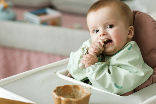 Cute little baby try to eat solid food in child chair. Complementary food. Adorable happy messy and funny infant portrait in porridge. Feeding after baby 6 months
