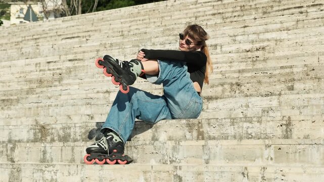 Gimbal orbital shot of young woman in wide jeans sitting on stone steps putting on orange-wheeled inline skates. Glances at camera, sunglasses, ponytail, knee pad. Warm sunny stone texture. Urban 