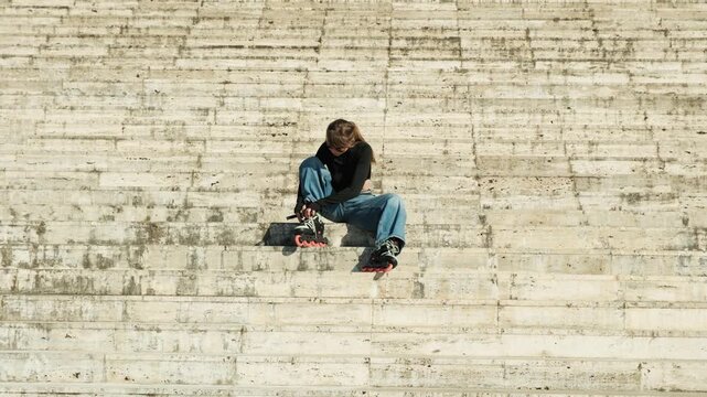 Gimbal pull back shot of young woman alone on vast stone steps putting on inline skates. Small figure against large textured staircase. Minimalist powerful composition, warm sunny light. Urban active 