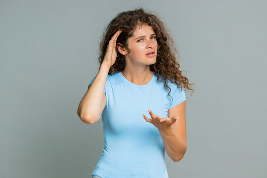 Young woman holding hand near ear gesture indicating difficulty hearing clearly, confused emotional reaction. Girl on gray background visibly expresses uncertainty asking to speak louder humorous mood