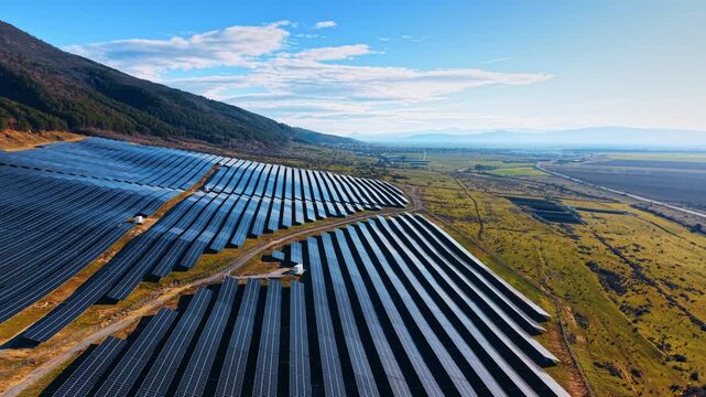 Solar panel arrays on hillside above rural valley. Aerial view of solar panel rows on sloped terrain with mountains, valley fields, clouds, and side light.