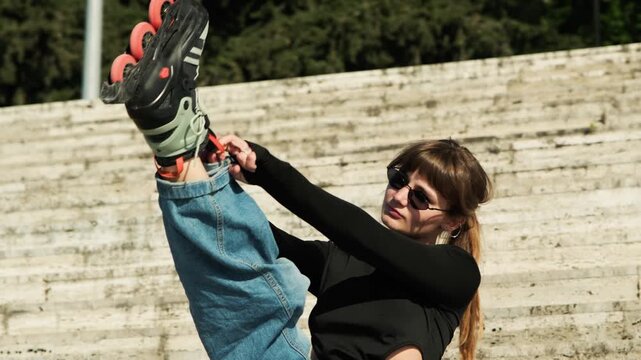 Gimbal orbital shot from medium to wide of young woman lifting leg high on stone steps to put on orange-wheeled inline skate. Cat-eye sunglasses, black cutout top. Minimalist stone staircase 