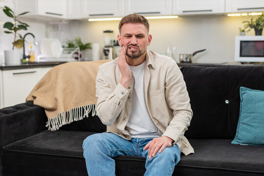 Young man at home gets sharp toothache covers cheek and grimaces then breathes slow for relief. Caucasian guy on sofa holds jaw with palm and shows pain worry and tense mood during hard wave and calm.