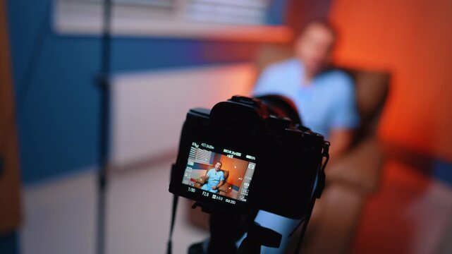 Camera Records Doctor Vlog In Clinic Interview Room. Foreground camera films doctor in chair, blurred clinic room, blue wall and orange side light.