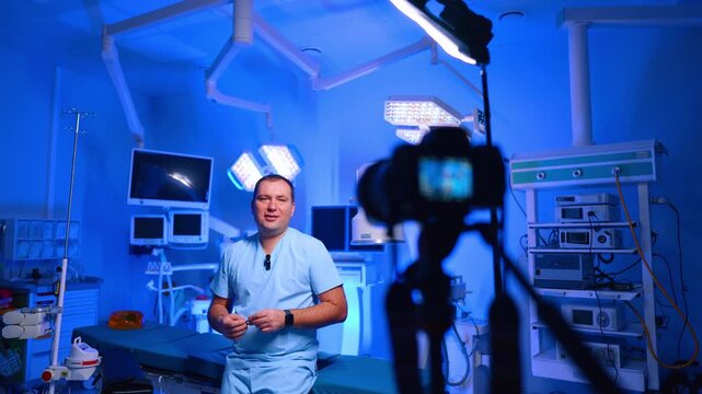 Doctor Films Medical Vlog In Blue Operating Room. Doctor in scrubs stands in operating room, camera silhouette foreground, blue lighting and surgical equipment.
