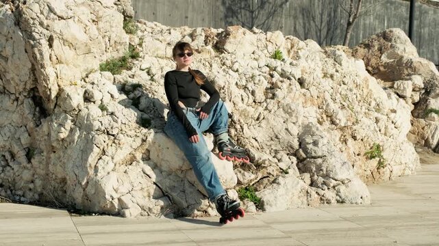 Gimbal orbital shot of young woman in wide jeans with orange-wheeled inline skates leaning against natural rocky cliff. Cat-eye sunglasses, hand on hip, confident pose. Sunny textured limestone