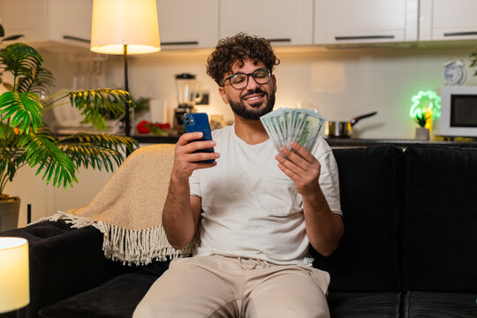 Arabian man at home counts dollar bills records salary sums in smartphone notes plans monthly budget. Egyptian guy on sofa reviews cash tracks expense lists goals for savings rent travel buys then up.