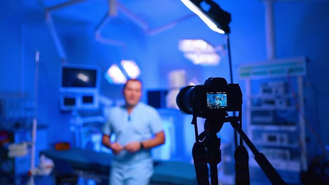 Camera Focuses On Doctor Filming Vlog In Operating Room. Camera on tripod in focus, doctor blurred behind, blue operating room and overhead LED light.