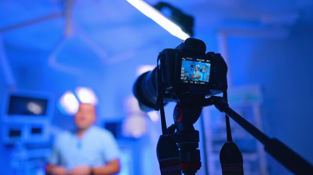 Tripod Camera Records Doctor Speaking In Blue Clinic. Tripod camera in foreground records doctor, blue clinic equipment and shallow focus background.