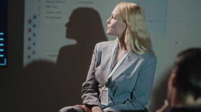 Tilt up shot of confident blonde Caucasian businesswoman sitting on chair in front of audience pointing at projection screen and giving speech during conference