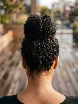 Natural curly hair top knot bun on a woman seen from behind in an outdoor setting at sunset. (96 karakter)