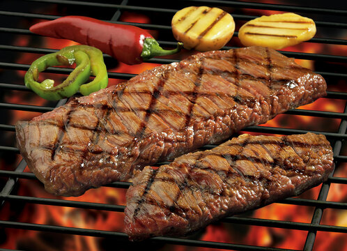 Grilled flank steak on a barbecue rack with flames, potatoes and peppers garnish