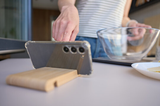 Smartphone stands on holder while person prepares food beside a glass bowl