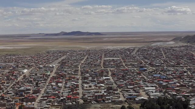 A view of Oruro overlooking the expansive Bolivian Altiplano. A wide-reaching plateau encircles the city, featuring brick houses topped with vibrant roofs beneath a pale, overcast sky.