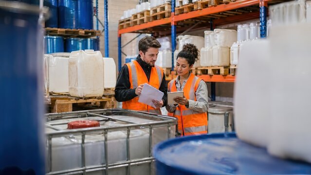 Male and female warehouse workers in safety vests checking inventory using a digital tablet and clipboard in an industrial storage facility with chemical barrels and IBC tanks.