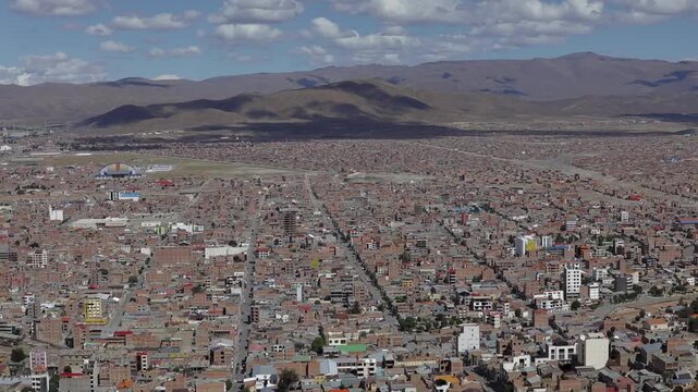 A high-angle view of Oruro, Bolivia, overlooking a dense urban landscape of unfinished brick houses. In the distance, the rugged Andean mountains frame the city skyline.