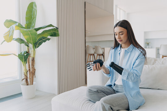 Young woman with prosthetic arm holding bionic hand while sitting on bed at home
