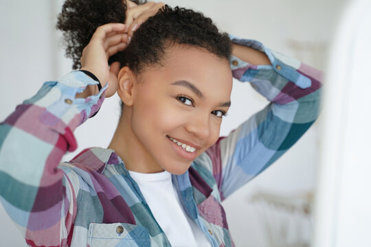 Young black woman tying curly hair and smiling in bright room at home