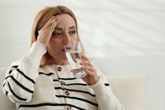 Menopause. Woman suffering from headache and drinking water on sofa at home. Space for text