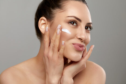Beauty woman portrait applying cream on cheek smiling, female model with glowing radiant skin demonstrating skincare routine, facial cosmetology and moisturizing beauty concept