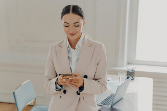 Business woman using smartphone in office by desk with laptop checking messages during workday