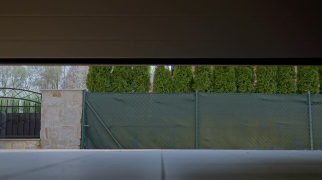 Garage door opens from an interior view to a fenced yard with trees, stone wall and exterior building. Concept of home access and security