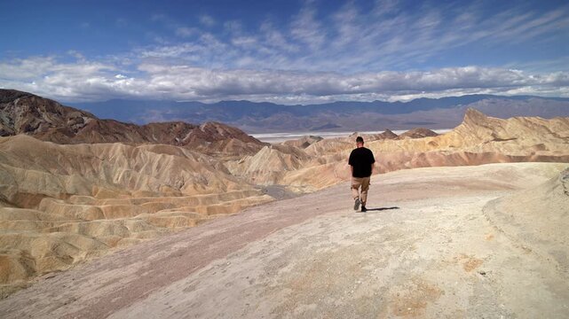Man wearing black t-shirt and khaki pants walks forward across barren desert terrain toward stunning golden and ochre colored eroded badlands of Zabriskie Point in Death Valley National Park.