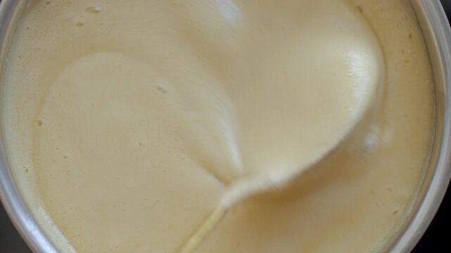 Spoon stirs light creamy mixture with foam bubbles in saucepan. Concept of cooking process and homemade recipe