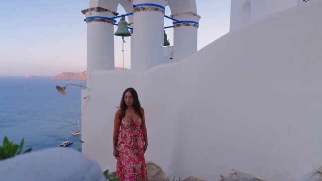 Beautiful woman wearing colorful floral maxi dress poses near traditional white Greek Orthodox church bell tower with scenic Aegean Sea and boats in background during golden hour. Santorini, Greece