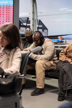 Man on holiday trip listening to music, waiting in airport lounge during delayed flights. African american person wearing audio headset waiting for cancelled flight to resume, sitting in terminal