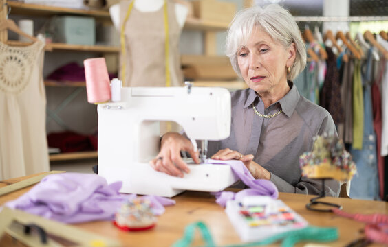 Skillful old tailoress sewing textile sitting at her workplace in tailor shop
