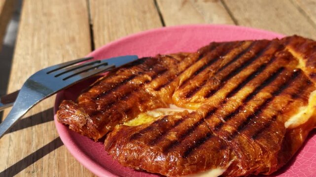 Grilled croissant planchado sandwich with ham and melted cheese on pink plate, woman girl cutting traditional breakfast pastry with knife and fork, crispy golden brunch food on wooden table, close up