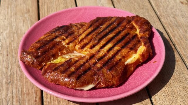 Grilled croissant planchado sandwich with ham and melted cheese on pink plate, woman girl cutting traditional breakfast pastry with knife and fork, crispy golden brunch food on wooden table, close up