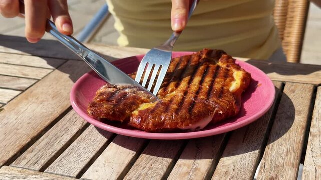 Grilled croissant planchado sandwich with ham and melted cheese on pink plate, woman girl cutting traditional breakfast pastry with knife and fork, crispy golden brunch food on wooden table, close up